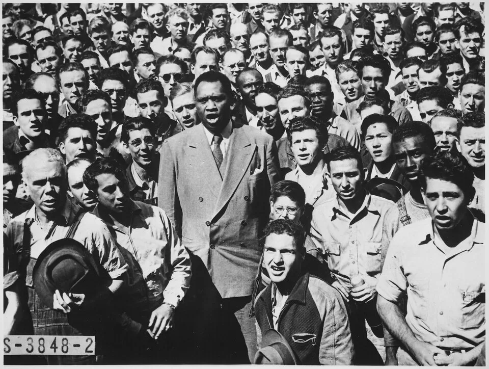 Black-and-white 1942 photograph of Paul Robeson in an overcoat, head raised and mouth open in song, surrounded by a dense crowd of shipyard workers in work clothes, several singing along with him.